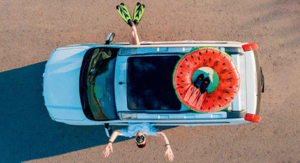 Vista aérea de un coche cargado con flotador de sandía y aletas de buceo, listo para las vacaciones de verano, con una persona tumbada al lado del vehículo.
